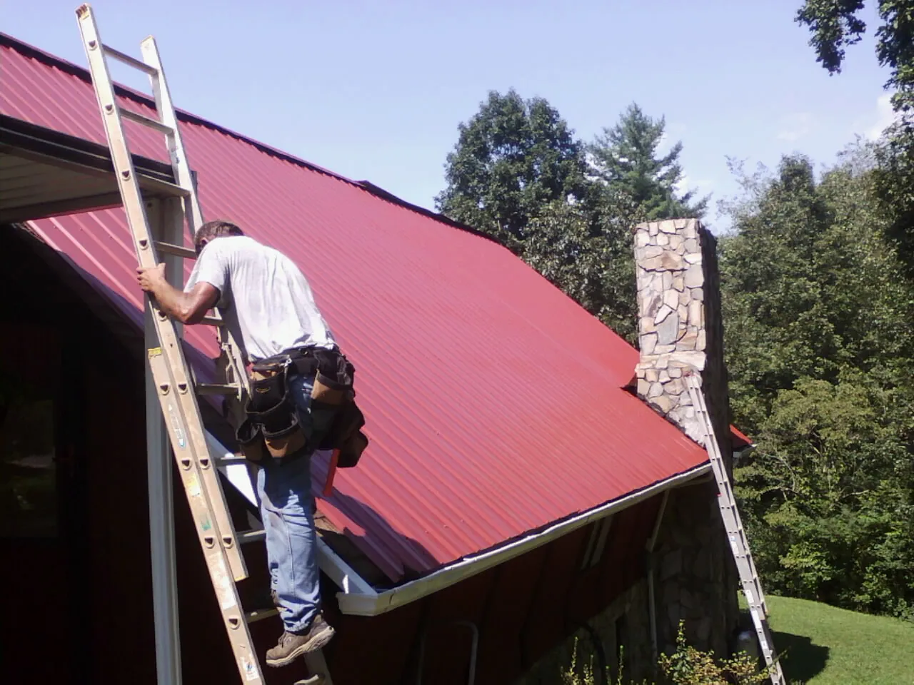 Roofer on a ladder beside a red metal roof and stone chimney — Soffit & Fascia Repair by Roofing Brothers in Texhoma